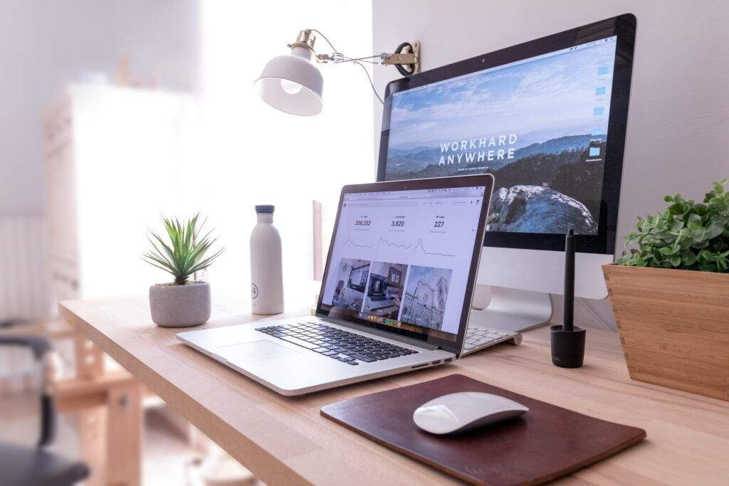 A minimalist home office desk setup with a MacBook and external monitor displaying analytics and a mountain landscape reading 'Work Hard Anywhere,' flanked by a small succulent, a white water bottle, a potted plant, and a wireless mouse on a leather desk pad.